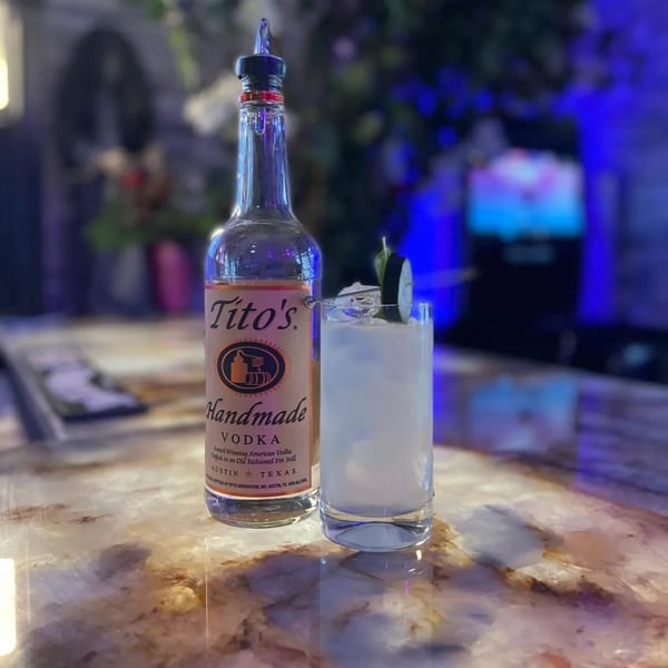 A Tito's Handmade Vodka bottle next to a cocktail garnished with cucumber on a marble bar.