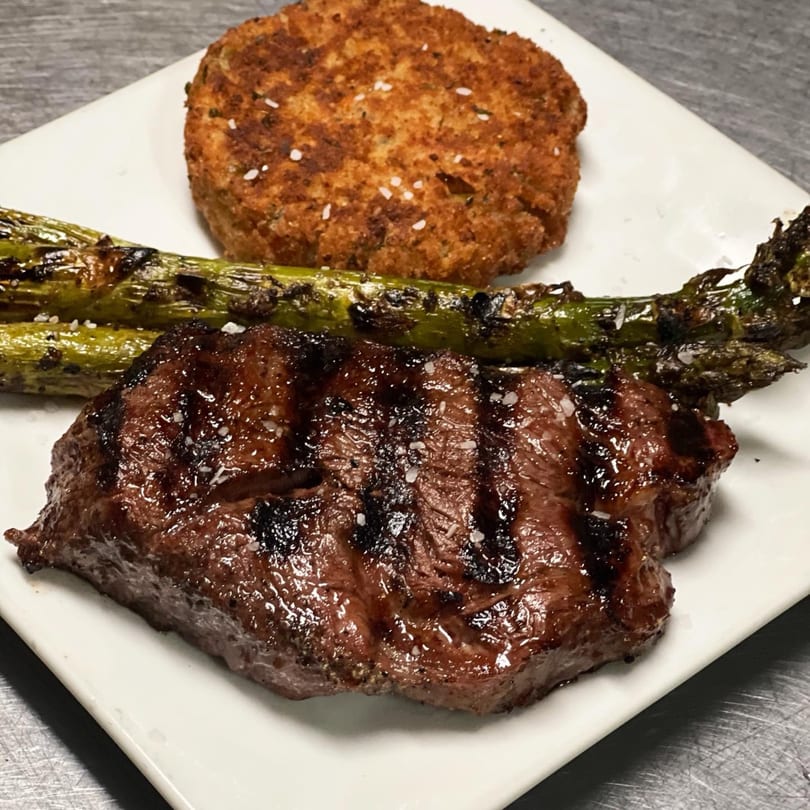 Grilled steak with grill marks, roasted asparagus, and a crispy crab cake on a white plate.