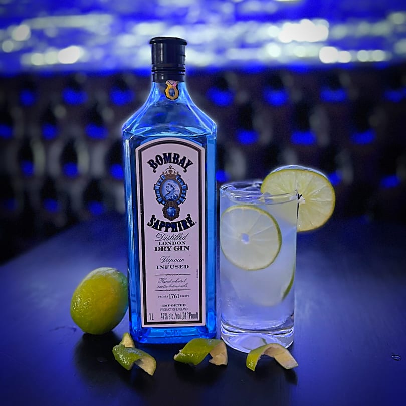 Bombay Sapphire gin bottle, a gin and tonic with lime, and fresh limes on a dark surface.