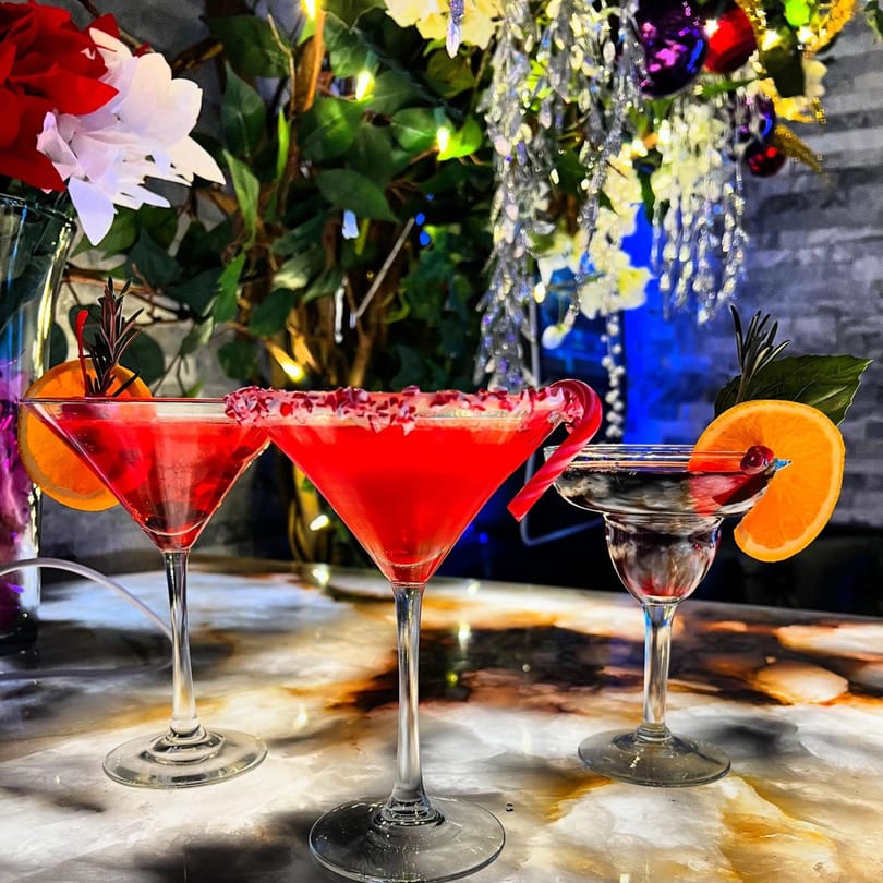 Three festive red cocktails, two martinis and one margarita, on a marble bar with holiday decorations.