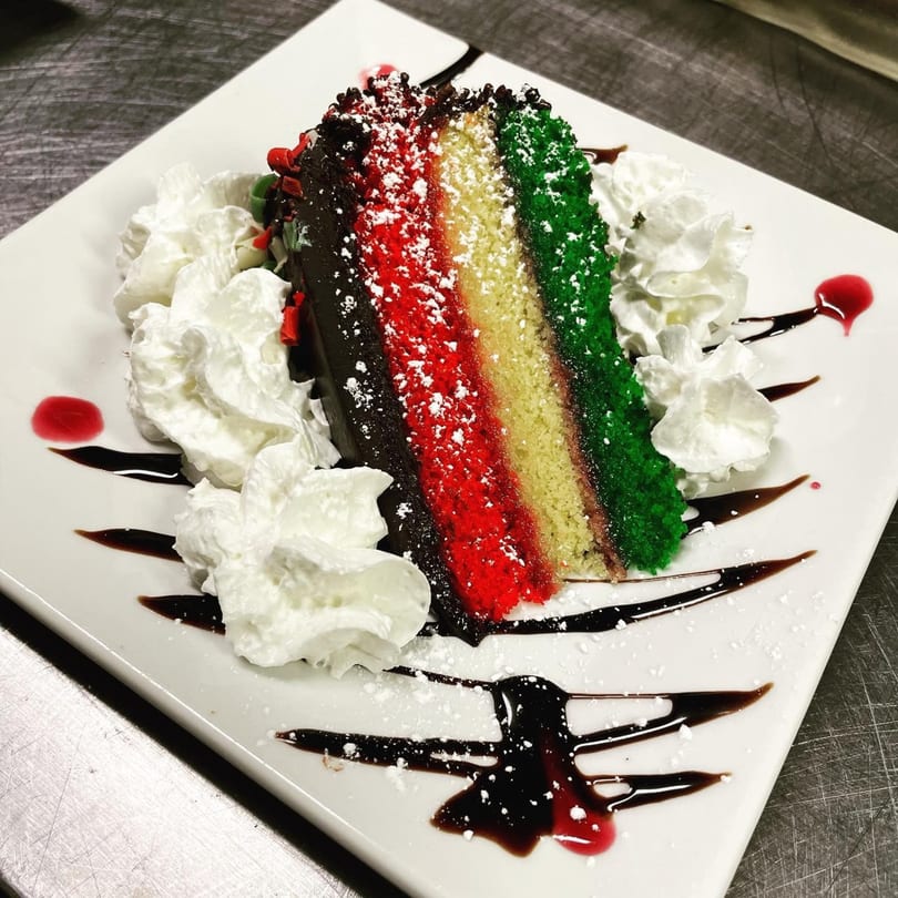 Slice of colorful layered cake with whipped cream, chocolate, and raspberry drizzle on a white plate.