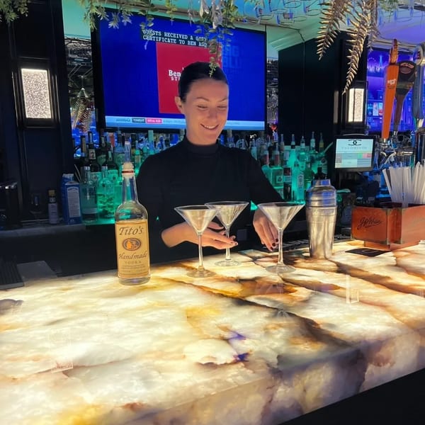 A bartender at Nunzi's American Bistro prepares drinks on a glowing marble bar, with Tito's vodka and martini glasses.