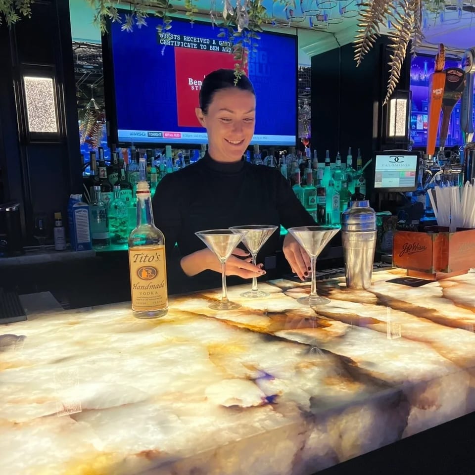 A bartender at Nunzi's American Bistro prepares drinks on a glowing marble bar, with Tito's vodka and martini glasses.