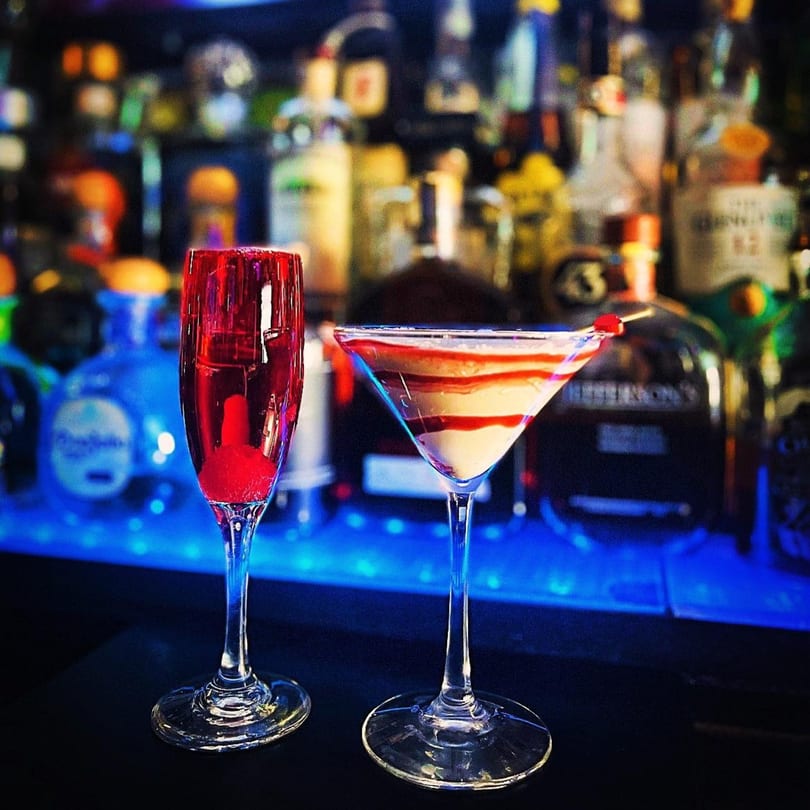Two cocktails, a red champagne flute and a striped martini, on a bar with blue lighting.