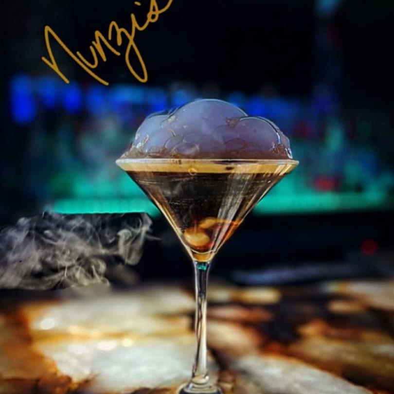A dark cocktail in a martini glass with a large smoke bubble on top and smoke swirling around the base.