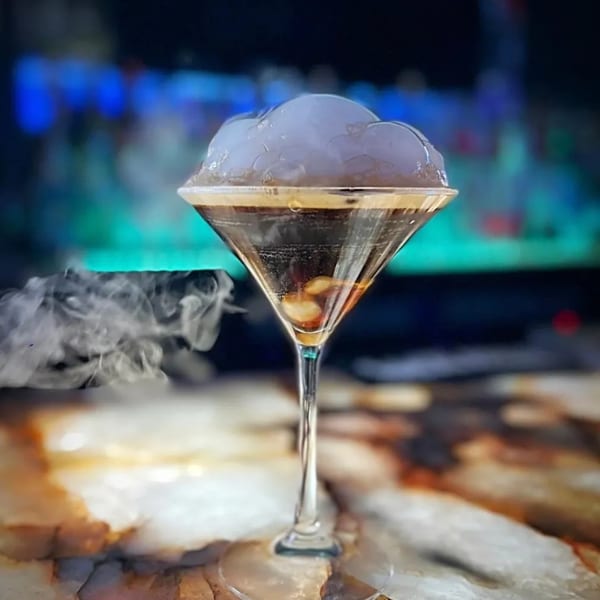 A martini glass with a dark drink, topped with a cloud of white foam and wisps of smoke.