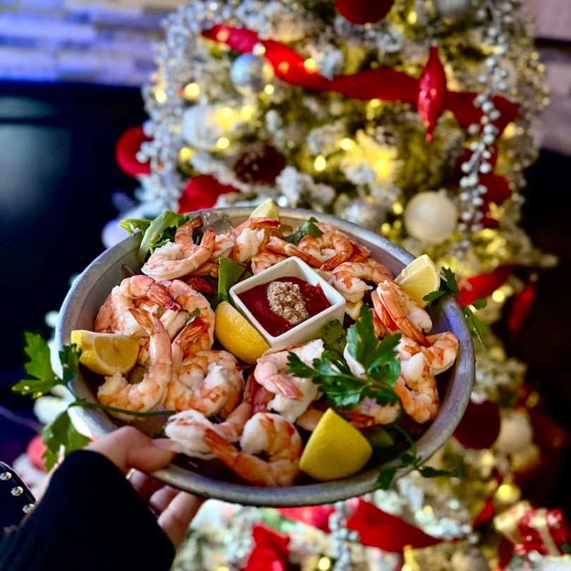 A hand holds a platter of shrimp cocktail with lemon wedges and sauce, in front of a festive Christmas tree.